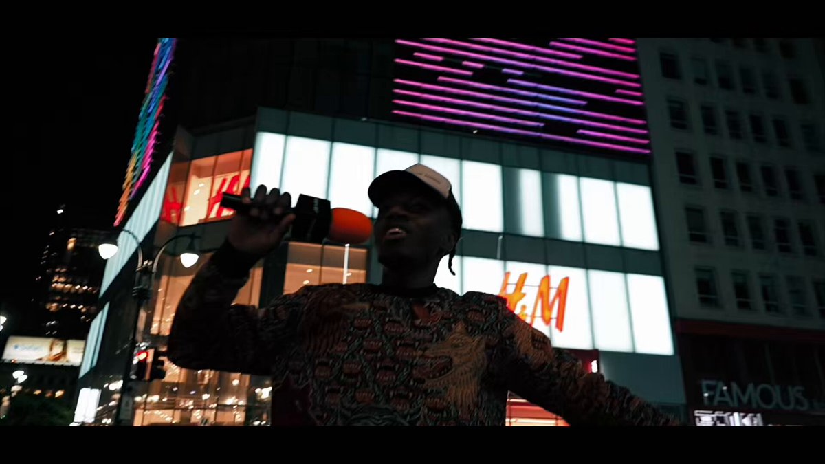 The image shows a man standing next to a building, holding a camera up to his face as if taking a picture of himself or someone else. The photo has been edited with a filter that makes him appear to be wearing sunglasses. The background features a cityscape at night, including buildings and neon lights. There is also an advertisement for H&M visible in the image.