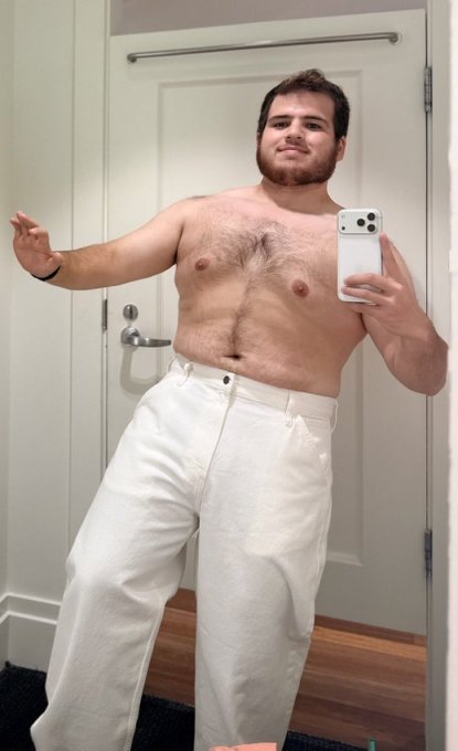 A shirtless man takes a mirror selfie wearing white pants, holding a smartphone to capture the image.