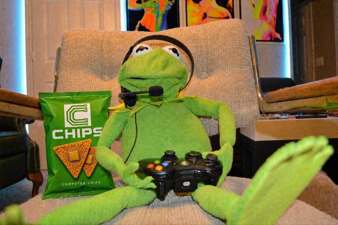 A Kermit the Frog plush toy sitting in a chair wearing headphones, holding a game controller, with a green bag labeled 'C CHIPS Computer Chips' beside it.