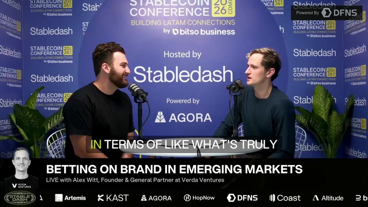 The image features two men sitting at a table with microphones behind them, likely participating in an interview or podcast discussion. The background of the image includes a blue wall adorned with various logos and text, such as "Stablecoin Conference", "Building Latam connections by OBOB Businesses", and "In terms of like what's truly". Additionally, there are several other texts present on the image that may be related to the interview or podcast.