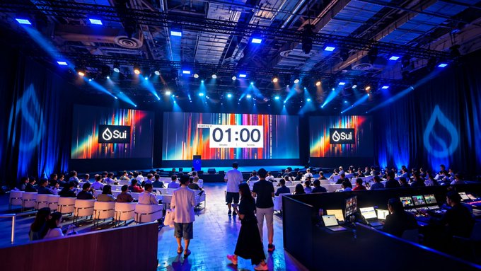 A large conference hall with an audience watching a screen displaying 'Sui' logos and a countdown timer at '01:00'.