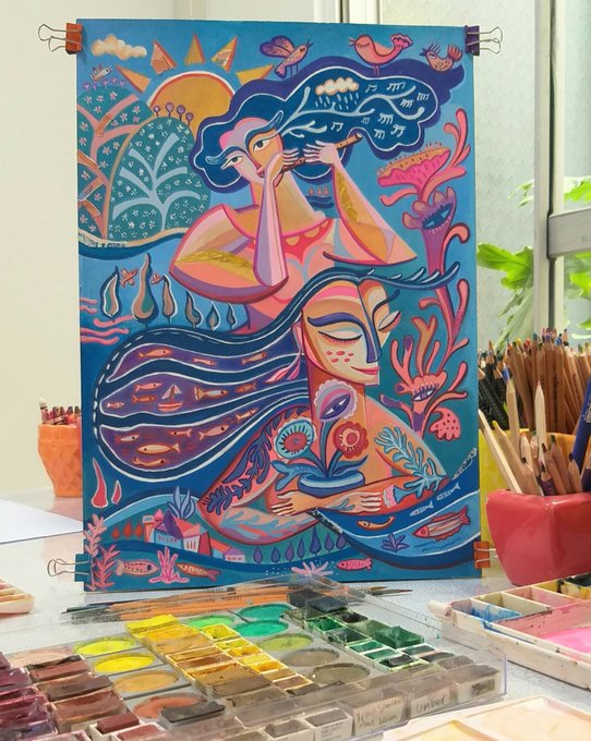 A vibrant, colorful painting on a canvas displayed on an art studio desk, featuring stylized faces, flowers, and birds in shades of pink, blue, and orange, surrounded by watercolor palettes and brushes.