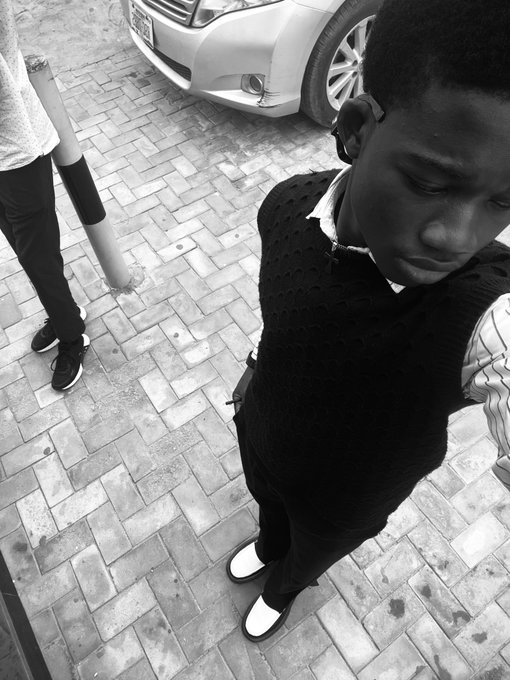 A black-and-white photo taken from above shows a young man in a dark sweater vest and white shoes looking down on a brick sidewalk, with a car and another person's legs visible nearby.