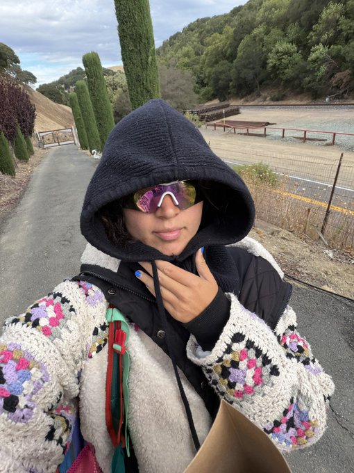 A person wearing a dark hood, reflective sunglasses, and a colorful crochet jacket takes a selfie on a path lined with cypress trees and hills in the background.