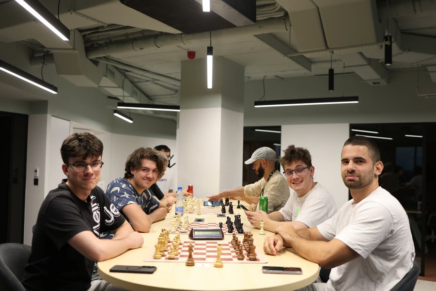 Six young men smile around a long boardroom table set up for a casual chess tournament, with multiple boards mid-play and a digital chess clock visible in the foreground. The scene captures the relaxed, communal atmosphere of the Yearnament chess event hosted with DeFiSaver at Eth Belgrade.