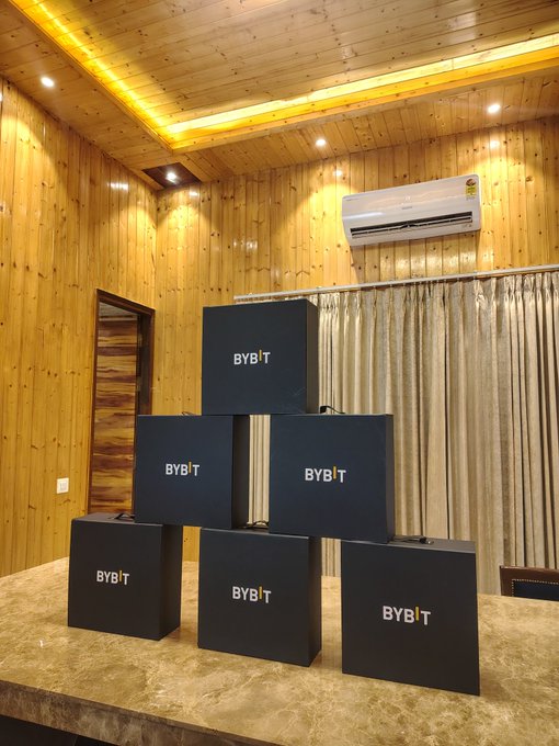 Six black Bybit branded boxes are stacked in a pyramid formation on a table in a wood-paneled room with a white air conditioning unit on the wall.