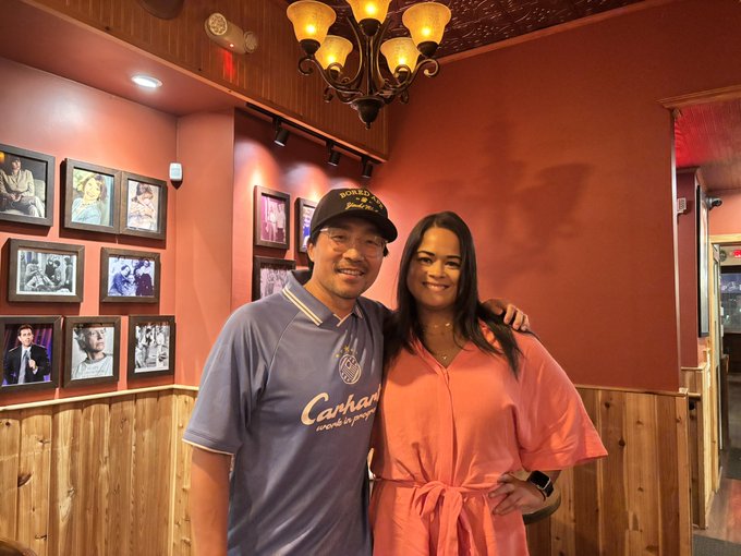 A smiling couple poses together inside a warmly lit restaurant with wood paneling, framed black-and-white celebrity photos on the walls, and a chandelier overhead; the man wears a blue 'Carhartt' polo and cap, while the woman wears a coral dress.