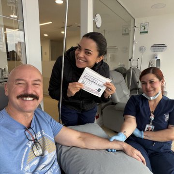 A bald man with a mustache sits smiling while donating blood, accompanied by a woman holding a certificate that reads 'COĞRAFYA' and a nurse in blue scrubs.