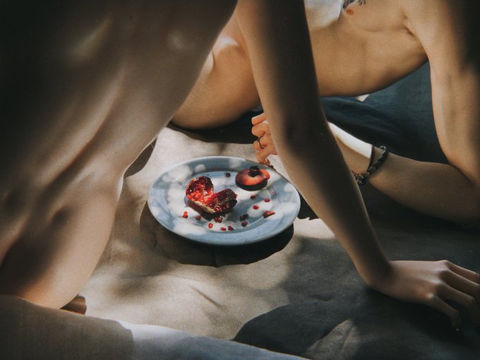 A person lies on a bed in soft light near a plate of fruit featuring a cut pomegranate, visually connecting to the 'arcadia. pomegranate' theme.