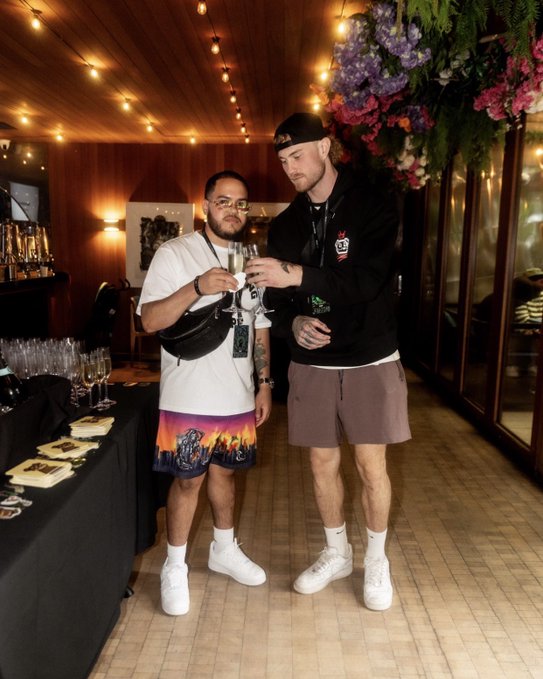 Two men stand together at an indoor event with floral ceiling decor and string lights, holding champagne glasses; one wears a white shirt with colorful shorts and a black crossbody bag, while the other wears a black hoodie, brown shorts, and a backward cap.