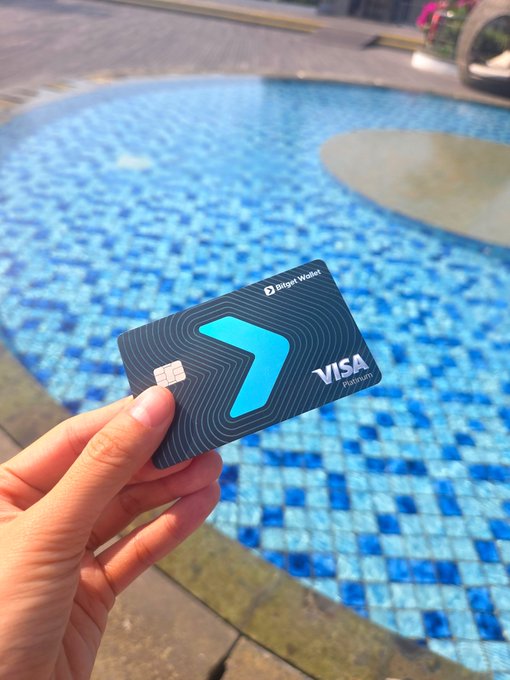 A hand is proudly displaying the Bitget Wallet VISA Platinum card in front of a tiled swimming pool, showcasing the sleek dark card with its signature blue logo as a visual promotion for Bitget's referral offer.