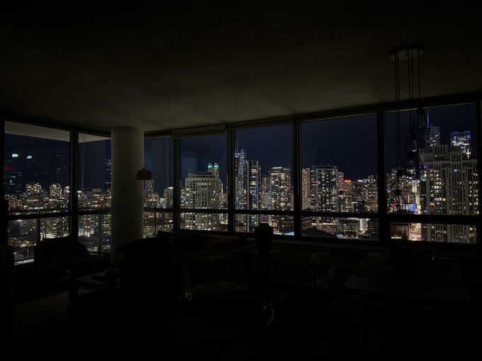 A dimly lit interior room featuring large floor-to-ceiling windows that offer a panoramic view of a city skyline illuminated at night.