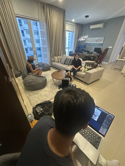 A man in the foreground works on a laptop displaying video editing software while two people relax on bean bags and a sofa in a modern living room.