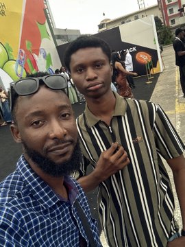 Two men pose together at what appears to be a Web3 or crypto event, with one taking a selfie while wearing sunglasses on his head and the other giving a peace sign in a striped shirt. Behind them, colorful murals and a banner with partially visible text reading "TRACK" can be seen, tying into the live Web3-focused post tagging crypto community members.