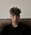 A young man with curly hair wearing a dark sweater, photographed in what appears to be a casual indoor setting with a couch behind him.