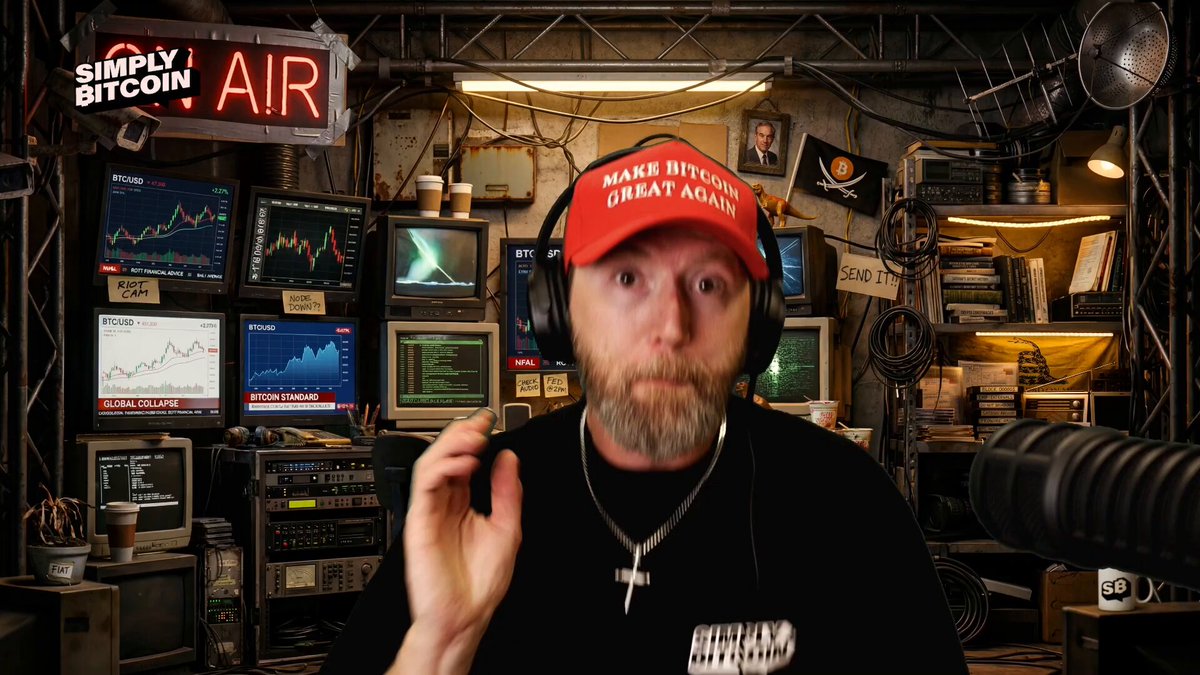 The image features a man wearing a red baseball cap, standing in front of a computer screen with a keyboard and mouse nearby. He appears to be engaged in an online meeting or video call while wearing headphones. The background includes several monitors displaying stock market data, indicating that he might be discussing Bitcoin or the cryptocurrency market.