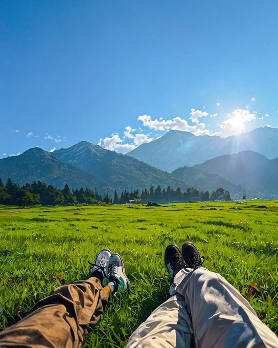 Two pairs of legs wearing sneakers rest on a grassy field, looking out toward a scenic mountain landscape under a bright blue sky.
