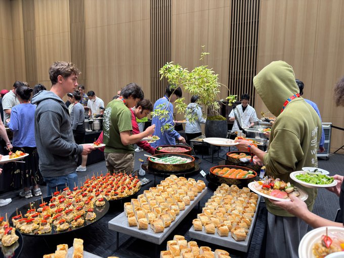 A crowded buffet scene where attendees serve themselves from a large spread of appetizers, including skewers, bread, fruit, and sushi, set in a modern room with wood-paneled walls.