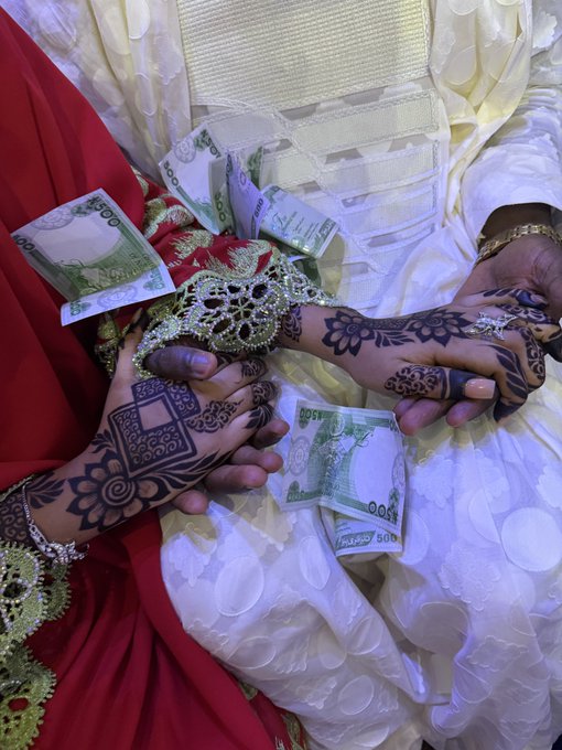 Two hands adorned with intricate henna designs rest against red and white traditional wedding attire, with several Saudi Riyal banknotes (including 500 and 100 denominations) tucked decoratively into the fabric, capturing a joyful moment of congratulations for @Gayu_BTC's wedding.