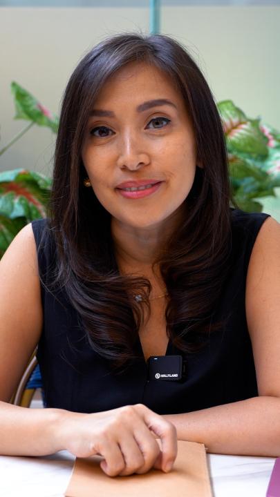 A portrait of a woman with long dark hair wearing a black sleeveless top and a small RØDE microphone, seated at a table with green plants in the background.