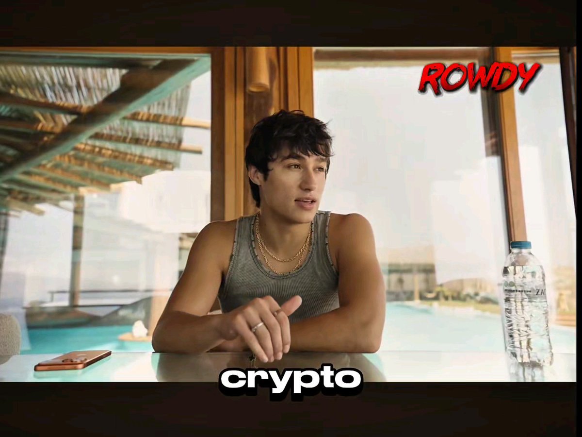 A young man in a gray tank top and gold chain sits at a table with a water bottle and phone, looking off to the side, while the image is overlaid with the text "crypto" at the bottom and "ROWDY" in red in the top right—visually representing the bullish crypto outlook mentioned in the post, where BTC at $60K is reframed as opportunity rather than a bear market.
