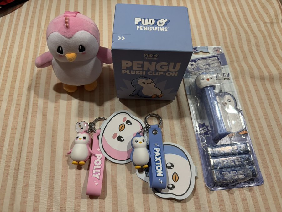 A collection of Pudgy Penguins merchandise including a pink plush toy, a 'PENGU PLUSH CLIP-ON' box, keychains labeled 'POLLY' and 'PAXTON', and PEZ dispensers.