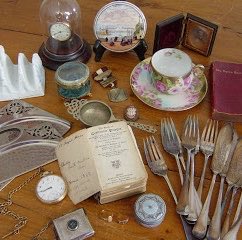 A collection of vintage heirlooms—including antique silverware, pocket watches, a clock, decorative china, and a small Bible with handwritten notes—is displayed alongside a modern smartphone, humorously illustrating the contrast between traditional family keepsakes passed down through generations and the digital devices millennials are likely to inherit.