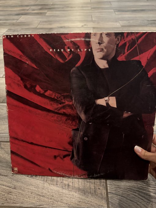 A hand holds up a worn vinyl record album of Tim Curry's "Read My Lips" against a wood-grain background, the album cover featuring the late actor in a dark coat against a dramatic red backdrop. The caption honors Tim Curry's passing alongside Dolly Parton, noting they'll be performing the "Time Warp" in tribute to his Rocky Horror legacy.