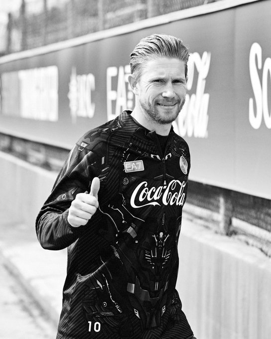 A black and white photo of a smiling man giving a thumbs up, wearing a dark patterned sports jersey with 'Coca-Cola' and 'EAT' sponsor logos and the number 10.
