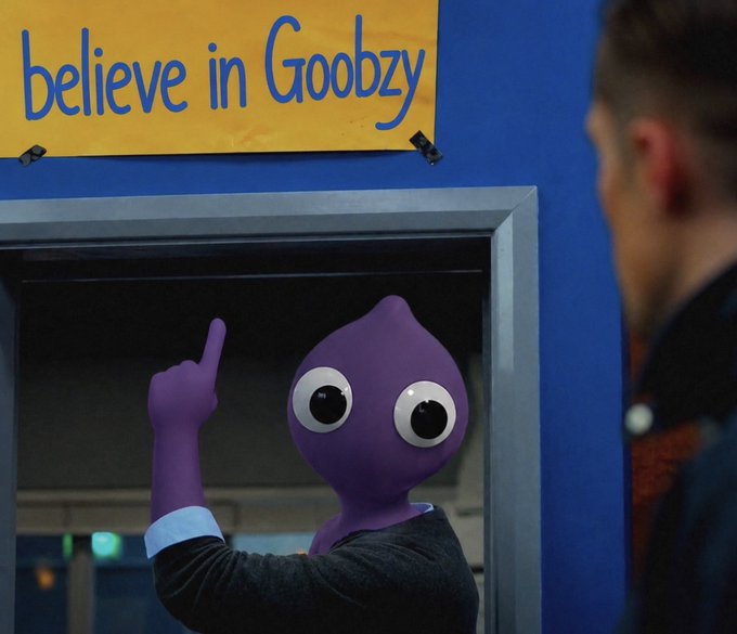 A purple mascot with large googly eyes points upward beneath a yellow "believe in Goobzy" banner, while a man in a denim jacket watches the scene unfold.