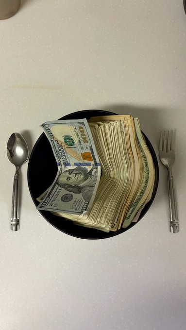 A black bowl sitting on a white surface filled with a thick stack of hundred-dollar bills, flanked by a silver spoon on the left and a silver fork on the right.