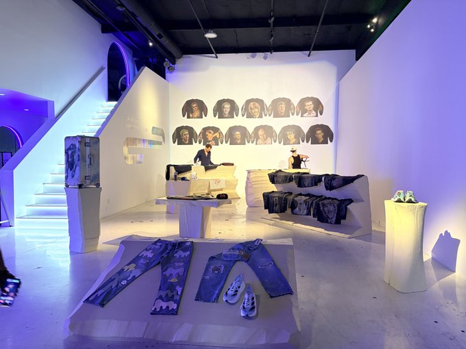 A brightly lit retail/exhibition space with purple and blue lighting displays merchandise on pedestals and mannequins, including clothing items with face graphics and footwear, set against a wall of repeated portrait prints.