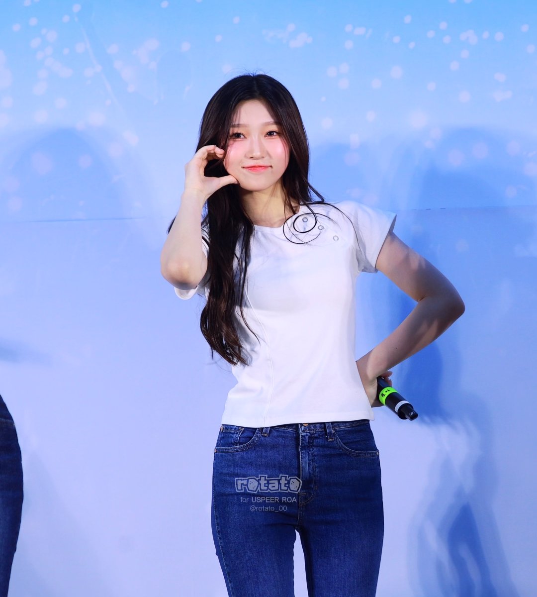 A young woman with long dark hair smiles while posing in a white t-shirt and blue jeans, holding a microphone with a green grip. Watermarks read 'rotato for USPEER ROA' and '@rotato_00'.