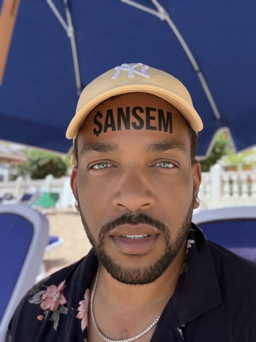 A selfie of a man wearing a tan NY Yankees cap and dark floral shirt under a blue umbrella at a beach, with the text '$ANSEM' edited across his forehead.