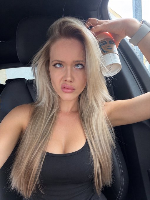 A blonde woman in a black tank top sits in a car, holding up a coffee cup with a face design on it.