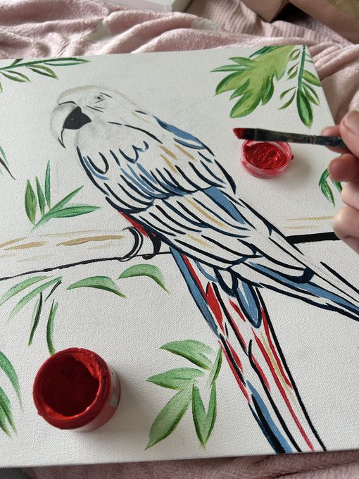 In-progress painting of a white and blue parrot on a branch with green leaves, with the artist holding a brush dipped in red paint.