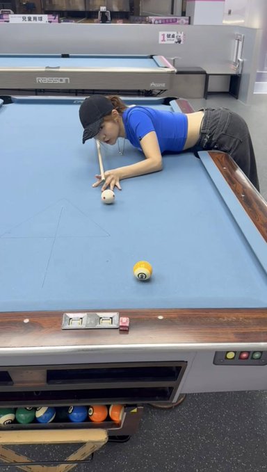 A woman wearing a black cap and blue crop top is playing pool, aiming her cue stick at the white cue ball on a blue felt table, with a yellow ball and a red cube visible.
