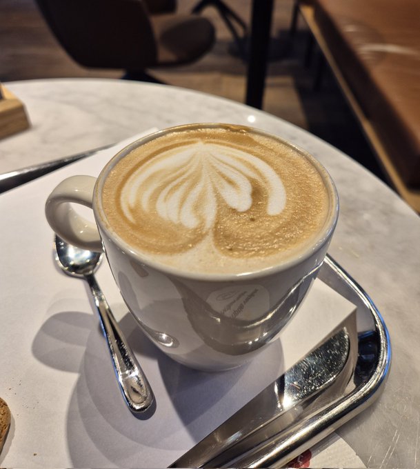 A cappuccino with intricate leaf-patterned latte art served in a white cup on a metal tray atop a marble café table, with a spoon placed beside it.