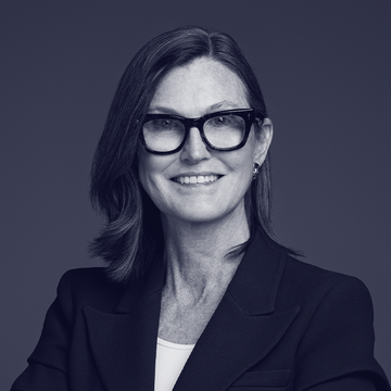 A professional portrait of a woman with shoulder-length hair wearing black-rimmed glasses and a dark blazer, smiling at the camera against a gray background.