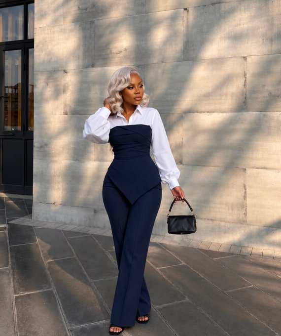 A woman with platinum blonde hair wears a white button-up shirt with a navy blue structured bustier and matching flared pants, posing on a sidewalk holding a black handbag.