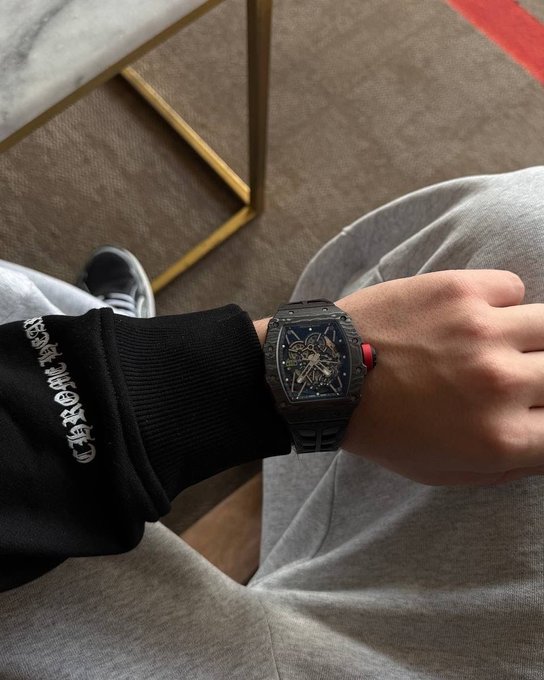 A close-up shot of a person's wrist wearing a dark, skeletonized luxury watch with a red accent button, paired with a black sweatshirt featuring white gothic-style text and gray sweatpants.