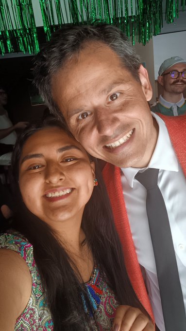 A smiling couple taking a selfie at an event; the man wears a white shirt, black tie, and red sash, while the woman has long dark hair and a colorful patterned top, with a person in glasses visible in the background.