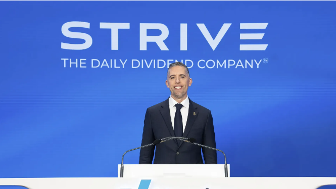 A man in a dark suit and tie smiling while speaking at a podium with a blue backdrop displaying the 'STRIVE - THE DAILY DIVIDEND COMPANY' logo.