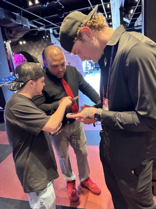 Three men are gathered together indoors at what appears to be an event, with one man in a black shirt and red shoes showing something on his smartphone to another man wearing a black cap, while a third man in a red tie looks on.