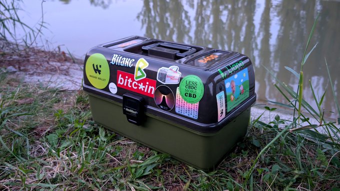 A green toolbox covered in stickers (including Bitcoin and various pop culture references) sitting in grass near a body of water.