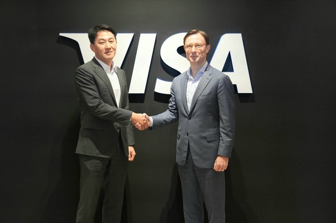 Two men in business suits shake hands in front of a large illuminated 'VISA' logo on a dark wall.