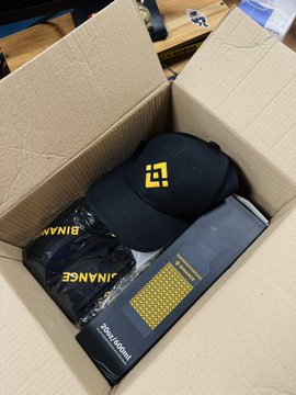 An open cardboard box containing Binance merchandise including a black cap with the yellow BNB logo, black shirts, and a colorful patterned item.