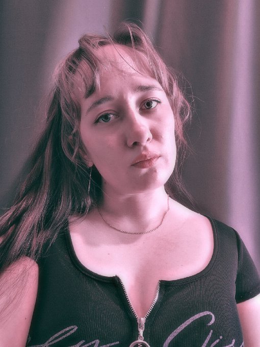 A woman with long brown hair and bangs, wearing a black zip-up top and delicate chain necklace, gazes directly at the camera against a soft curtain backdrop bathed in pink lighting. Her contemplative expression fits the morning greeting shared with the #NFTCommunity.