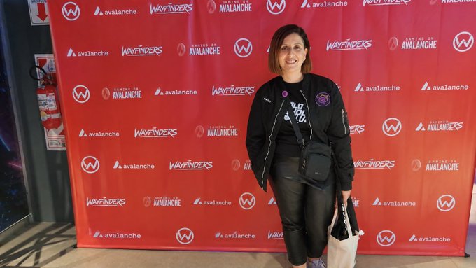 A woman in a black jacket stands smiling in front of a red branded backdrop featuring logos for 'Avalanche' and 'Wayfinders.'