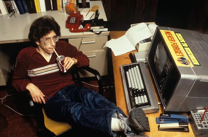A young man in glasses relaxes with a Coca-Cola while using an early desktop computer featuring "US Air Force" and "Navy" stickers, with papers and office supplies scattered around. The retro tech aesthetic reinforces the post's message about outdated trading approaches, suggesting traders should ditch "hard mode" leveraged strategies for more sustainable platforms like FX100Perp.